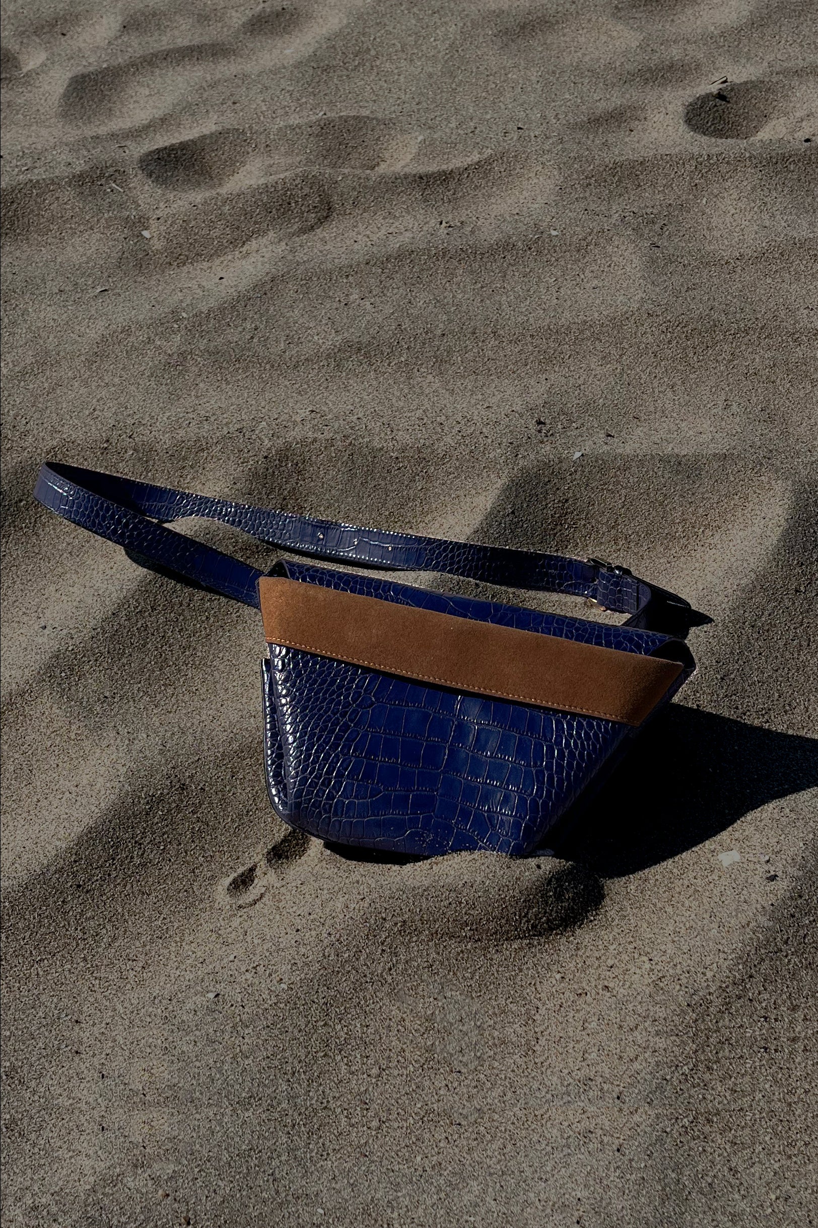 The Carrier Leather Cross Shoulder Bag in color Navy Embossed Croc resting on the beach.